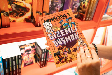 Naklejka premium 09 July 2024, Istanbul, Turkey: A woman in a store, examining the cover of a Minecraft-themed novel, surrounded by merchandise