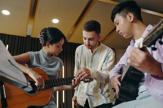 Music instructor teaching guitar techniques to two young students. The students are holding guitars and watching the instructor closely in a well-lit modern classroom setting