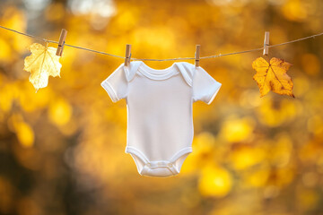 White baby onesie hanging with two yellow leaves on a clothesline outdoors in autumn background with copy space, fall baby pregnancy announcement	