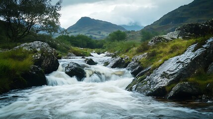 A river flows through a rocky valley with green hills in the background. The water is rushing and white and the rocks are covered in moss.