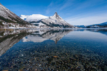 Lush mountains tower majestically over a tranquil lake, where the icy peaks and clear blue sky are...