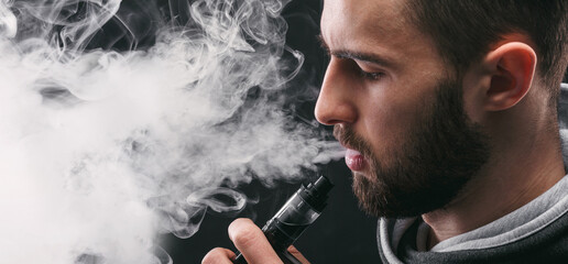 Man with vaping mod exhaling steam at black studio background. Bearded guy smoking e-cigarette to...