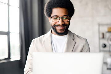 Charismatic Brazilian businessman focuses on his laptop screen, his friendly demeanor, underscored by a stylish and minimalistic office backdrop, portrays a blend of professionalism and accessibility