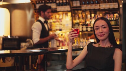 Beautiful guest looking at camera drinking at luxurious counter bar in mixed vodka Cosmopolitan red cocktail decorated with flower on bartender background in perfect atmosphere at nightclub. Vinosity.