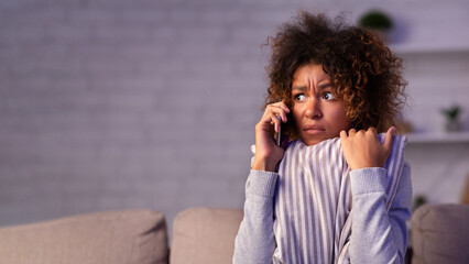 Scared afro woman calling phone and embracing pillow on sofa, afraid of noise near house, free space