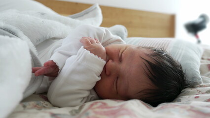 Newborn with dark hair sleeping peacefully on a bed, hands gently resting near the face, surrounded by soft blankets, showcasing the calm and serene nature of a baby's early life moments