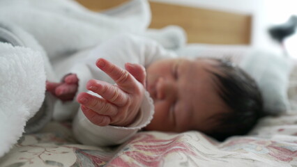 Newborn with dark hair sleeping peacefully on a bed, hands gently resting near the face, surrounded by soft blankets, showcasing the calm and serene nature of a baby's early life moments