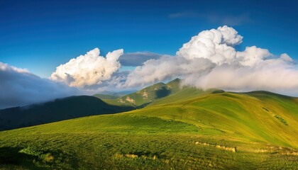Obraz premium Sunlit Green Hills with Rolling Clouds Overhead on a Clear Blue Sky Day, Capturing the Serene Beauty of a Misty Mountain Landscape with Vivid Grass and Dramatic Light Play in the Early Morning