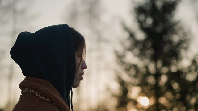A melancholic girl walks through a park at sunset, wearing a brown coat and a hoodie with the hood up, partially concealing her face. blurred background of trees and fading sunlight