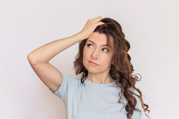 Fototapeta premium Young beautiful brunette woman with a curly hair and wearing a blue t-shirt looks thoughtfully to the side, puzzled by something, holding her hand on her head