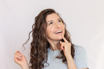 Young caucasian pretty brunette woman with a curly hair and wearing a blue t-shirt looks up, dreaming, smiling and holding a strand of hair in her hand. Isolated on a light background. 