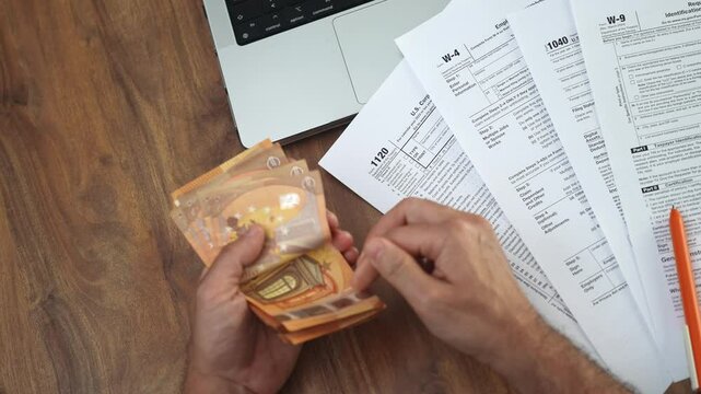 Self-employed man attentively unfolds several euro bills in hands calculating total sum at wooden table with collection of tax documents and laptop slow motion