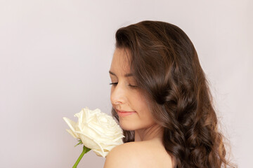 Young beautiful Caucasian brunette woman with curly hair smiles and with pleasure gently sniffs a white rose