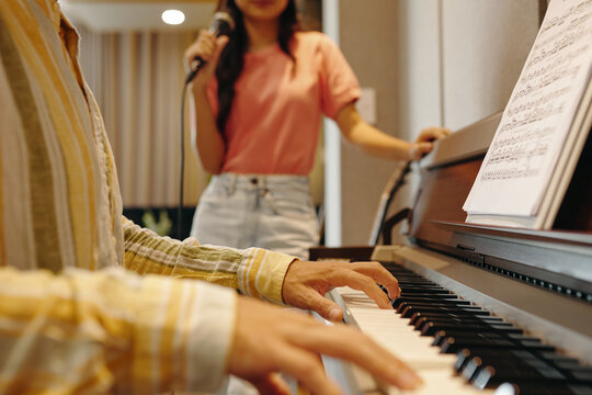 Person playing piano with sheet music while another person in background singing into microphone, capturing ambiance of music practice. Fingers on piano keys and focus on hands