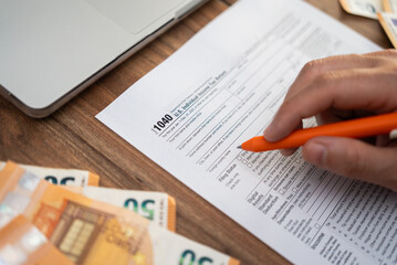 Obraz premium Close-up of a hand filling out a 1040 tax form with euros and a laptop on a wooden desk. Tax filing and financial preparation concept, ideal for accounting and international finance themes 