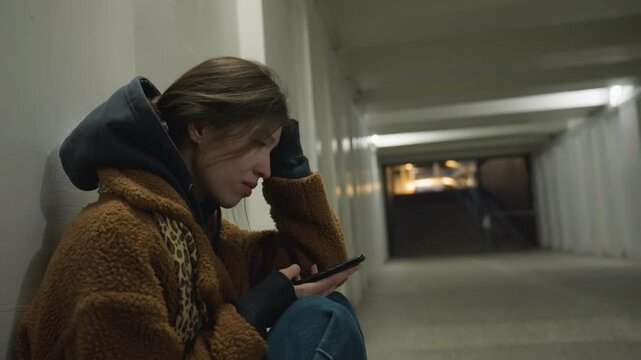 A close-up shot of a girl in a brown coat, sitting bent over in an underpass tunnel, holding her phone with a tearful expression. The scene captures an emotionally charged moment of sadness