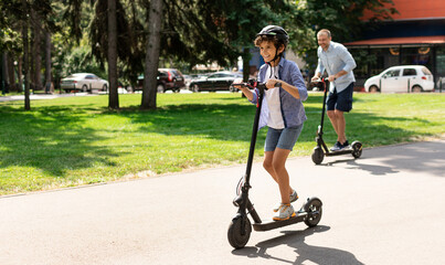 Happy Loving Family. Full body length photo of excited curly boy in protective helmet driving motorized push scooter with daddy at park, spending time together outdoors at summer, selective focus