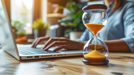 A modern hourglass filled with golden sand is placed beside a laptop, with a person typing in a bright and airy workspace, capturing a moment of focused work.