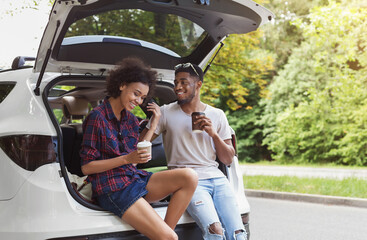 Happy afrocouple sitting in open car back, having stop off, drinking coffee. Romantic traveling concept