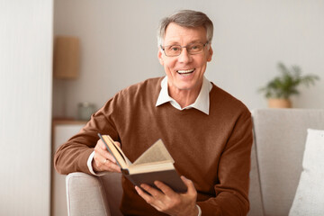 Positive Senior Man Holding Book Smiling At Camera Sitting On Sofa Indoor. Reading And Self-Development. Selective Focus