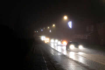 Car coming towards and past viewer along a dark misty night time urban road with headlights
