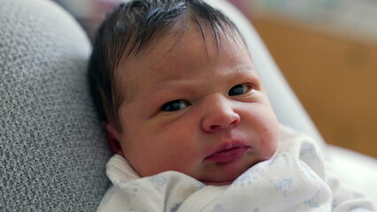 Close-up of a newborn baby with dark hair, displaying a curious and slightly furrowed brow. The...