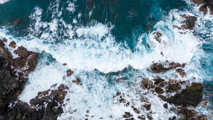 Aerial view of crashing ocean waves against rugged coastal rocks, displaying the dynamic beauty of turbulent sea foam and rocky shoreline.