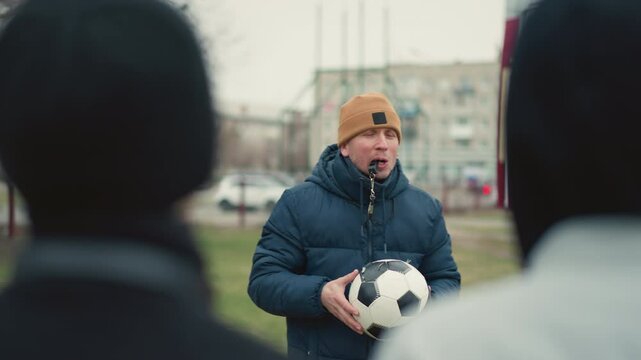 Close-up of a coach instructing two boys with a whistle in his mouth, holding a soccer ball, the coach throws the ball at the person wearing a gray top