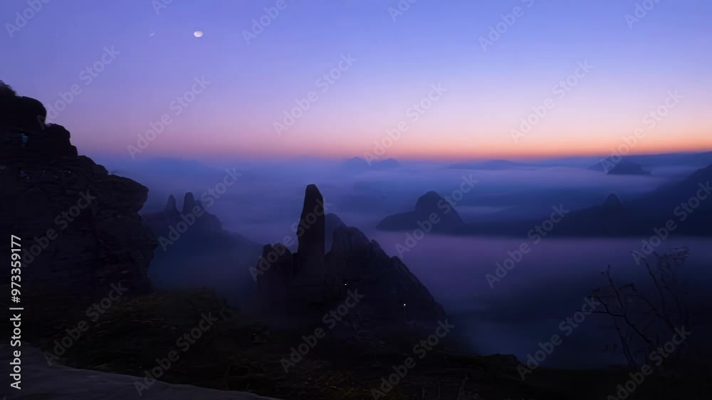 Poster The last hues of twilight are fading from the sky as the moon shines over the peaks of a mountain range emerging from the fog