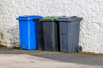 Blue, black, grey UK wheelie waste disposal bins standing beside white wall bin storage area in yard