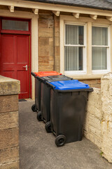 Grey UK wheelie wheeled waste disposal bins with grey, blue, red lids in bin storage area in front...