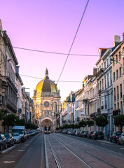 Naklejka premium A city street at sunset with buildings, a dome, tram lines, and parked cars. The scene highlights the urban design and architecture, creating a picturesque and vibrant atmosphere.