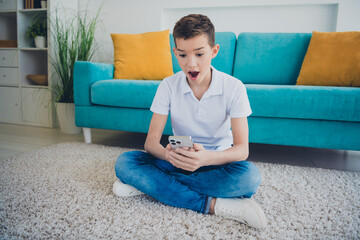 Full size photo of cute schoolboy sit floor use phone wear t-shirt modern interior apartment indoors