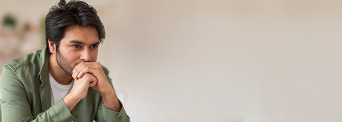 A young Indian man sits in a well-lit room, deep in thought with his hands clasped, indicating...