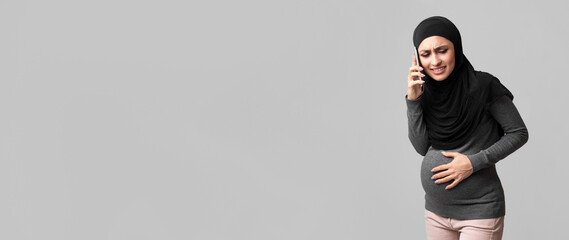 Pregnancy Pain Concept. Pregnant muslim woman in hijab suffering from abdominal cramps, calling to doctor for consultation and help, grey background