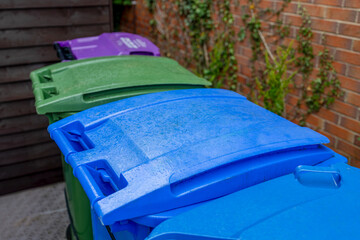 Plastic Waste Recycling Wheelie Bins lined up and secured in a UK garden storage area beside house brick wall, waiting for weekly council collection