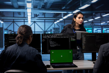 Multiethnic team in server hub ensures data remains shielded from potential threats using green screen laptop. Diverse colleagues safeguarding data center against malicious access with mockup notebook
