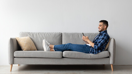 Obraz premium Cool Gadget And Application. Portrait of young smiling Arab man holding mobile phone, typing sms message, sitting on the sofa in living room. Guy browsing internet, surfing web, using app, free space