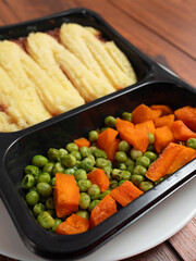 Cottage pie meal with peas and carrots in dark plastic container from microwave on white plate and wooden table. High quality meal for quick dinner at office or home. Popular British style food.