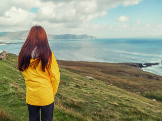 Teenager girl looking at amazing Irish nature scenery with ocean and mountains of Achill island, Ireland. Travel and tourism. Popular travel and tourism area with stunning view. Warm day, cloudy sky.