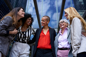 Low angle of team of diverse business only women laughing together happy at entrance of modern office block. Cheerful professional female group embracing each other in formal attire gathered outdoors