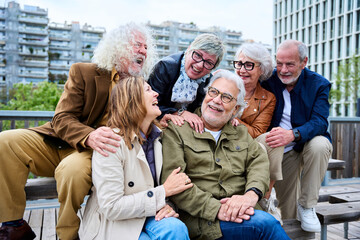 Group of six seniors grey hair joyful people having fun together sitting on bench in urban park. Cheerful mature Caucasian friends meeting talking funny enjoying free time laughing autumn day outdoors