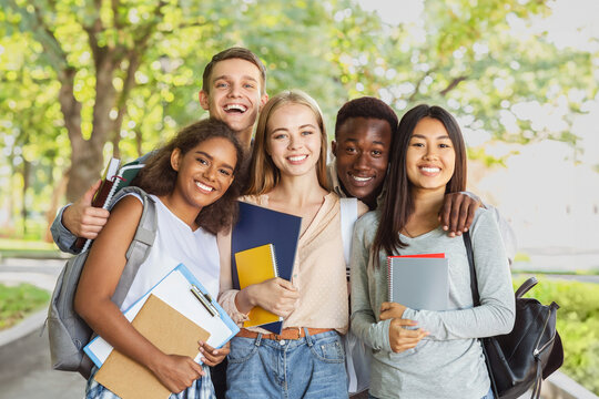 Group of international happy students with books and notebooks having fun in park after studying, smiling at camera