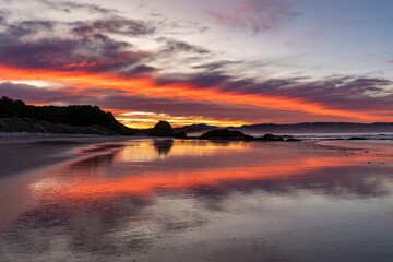 Beach at sunset with vivid firey reflections from the sun and clouds. Tawharanui, Warkworth, Auckland, New Zealand.