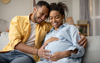 Pregnancy. Black Husband And Pregnant Wife Hugging Sitting At Home. Family Couple Expecting Baby, Man Embracing Woman Smiling Posing Together. Childbirth Concept