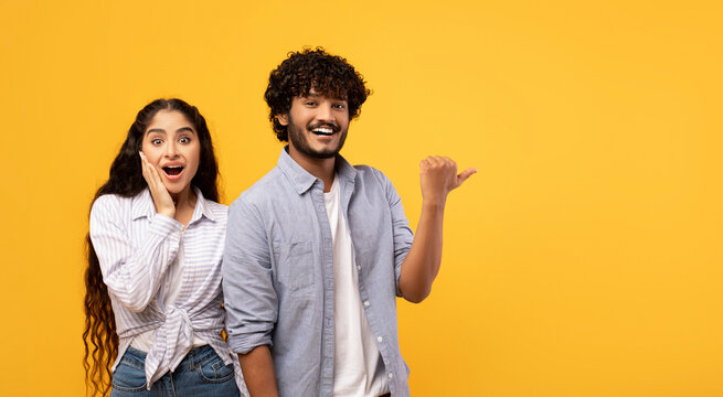 Omg, check this. Emotional indian guy pointing finger aside at free space over yellow background, surprised lady looking at camera, panorama. Couple demonstrating place for your text