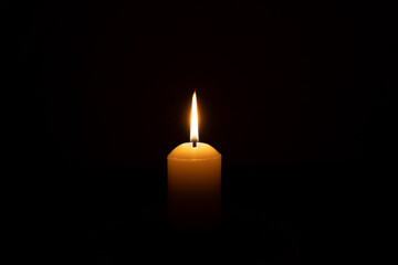 A single burning candle flame or light glowing on a big white candle isolated on black or dark background on table in church for Christmas, funeral or memorial service