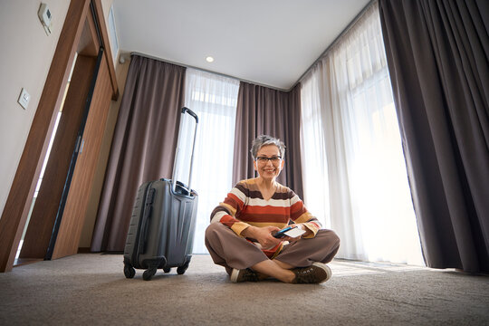Happy adult woman sitting in lotus position on the floor in hotel room, holding tickets