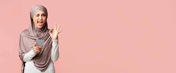 Cool islamic app. Positive muslim girl in hijab winking and showing ok gesture while using smartphone, standing over pink background with copy space