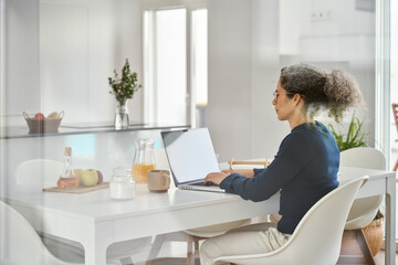 Busy middle aged woman sitting at table at home and using laptop in modern house. Mature 50 years...